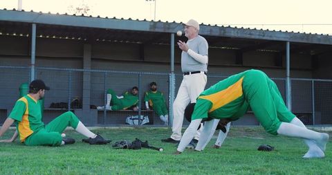 Senior coach leading pregame warmup, players stretching tossing baseball near dugout