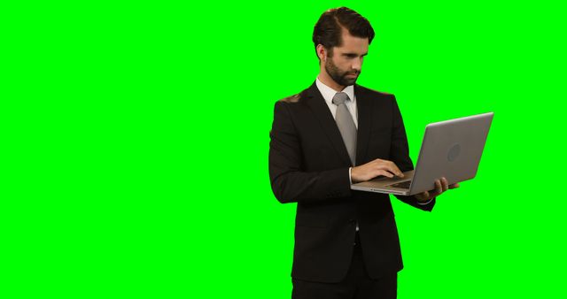 Businessman dressed in formal suit working on a laptop against a green screen background. This versatile image is perfect for creating business-themed presentations, corporate websites, advertising, and promotional materials where a professional look is required. The green screen allows for easy editing and customization.