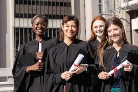 Multiracial female graduates celebrating outdoors holding diplomas and smiling together