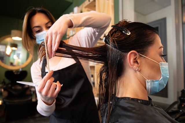 Hairdresser cutting hair of female customer in salon, both wearing face masks for safety. Ideal for illustrating health and hygiene practices in workplaces during the Covid-19 pandemic. Useful for articles on beauty services, professional hairstyling, and safety measures in salons.