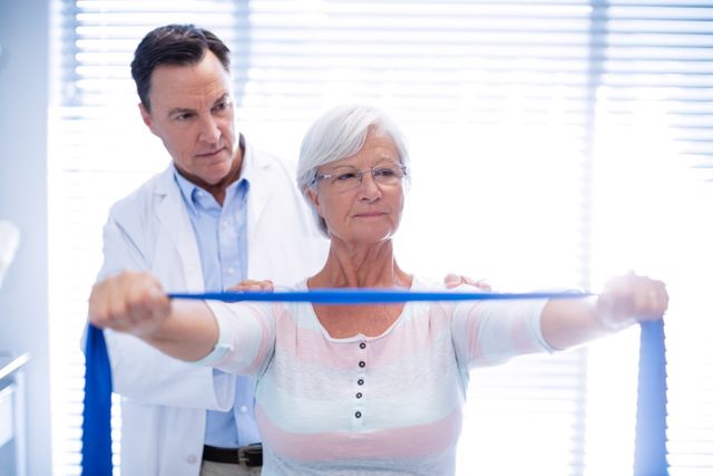 Senior woman receiving physiotherapy using a resistance band under the guidance of a male physiotherapist in a clinic. Ideal for illustrating concepts of elderly care, physical rehabilitation, healthcare services, and senior fitness programs.