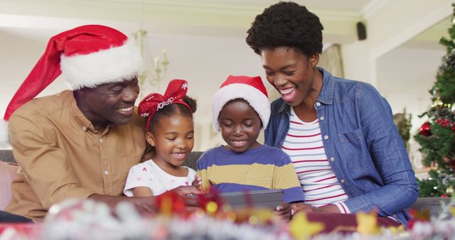 African American family sitting together at home during Christmas, all wearing Santa hats and looking joyful. Ideal for use in holiday advertisements, family-themed social media posts, and festive greeting cards emphasizing family togetherness and holiday cheer.