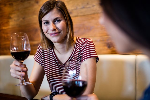 Young woman smiling and holding a glass of red wine while socializing with a friend in a bar. Ideal for use in advertisements for bars, restaurants, social events, or lifestyle blogs focusing on friendship and leisure activities.