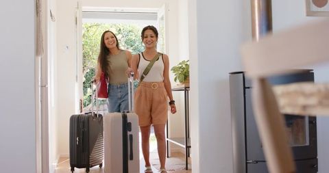 Two women walking into sunlit entryway pulling suitcases, smiling, summer travel