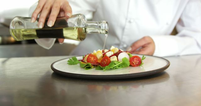 Chef in white coat adding olive oil to a fresh salad with tomatoes, herbs, and vegetables on a plate. Ideal for topics related to culinary arts, healthy eating, restaurant marketing, food blogs, and fine dining.