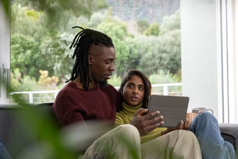 Diverse couple relaxing on sofa with tablet and mug by sunlit balcony in cozy home