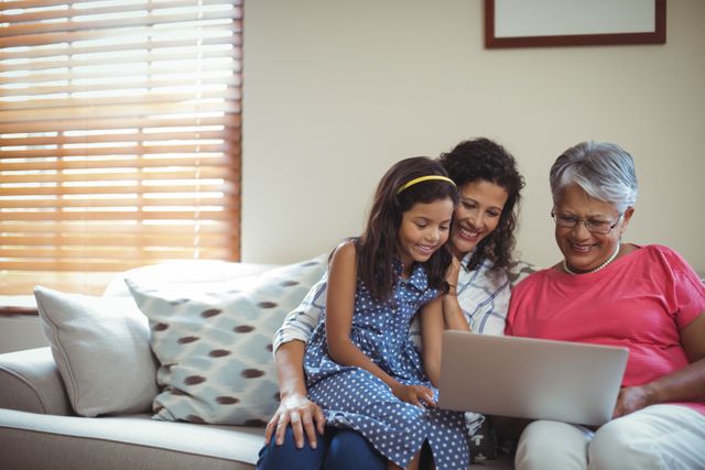This image shows a happy multigenerational family, including a grandmother, mother, and daughter, using a laptop together in a cozy living room. Ideal for use in advertisements, articles, or blog posts about family bonding, technology use in households, or multigenerational living. It can also be used for promoting family-friendly products or services.
