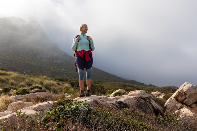 Senior woman with backpack standing on rocky terrain in mountainous area, surrounded by fog and greenery. Ideal for promoting active lifestyle, outdoor adventures, senior fitness, and travel destinations.