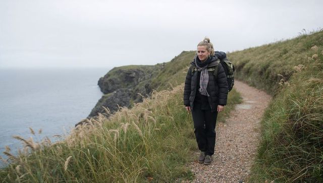 Solitary coastal hiker walking gravel cliff path with backpack in moody overcast seascape