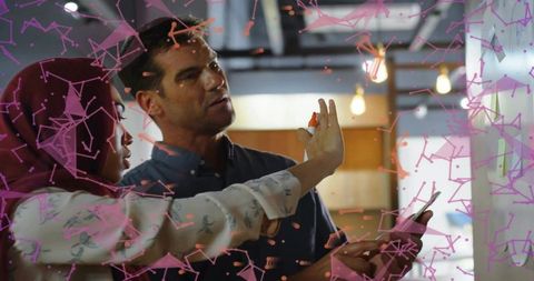 Diverse pair collaborating on glass board, hijab woman marking ideas while man gesturing