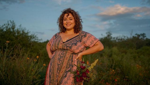 Curly-haired woman posing in wildflower meadow at golden hour holding red bouquet smiling