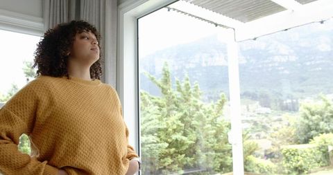 Curly-haired woman gazing out window at mountain view wearing mustard sweater
