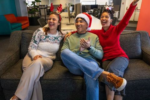 Diverse coworkers celebrating office holiday on sofa wearing santa hat, reindeer antlers
