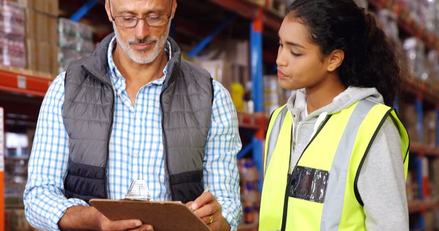 Warehouse supervisor providing instruction to a young employee on an inventory check. They are standing in a warehouse environment, both focused on a clipboard. The supervisor is wearing a checkered shirt and vest, while the employee is wearing a high-visibility safety vest. This could be used for themes related to teamwork, training, logistics, warehousing, inventory management, and mentorship in the workplace.