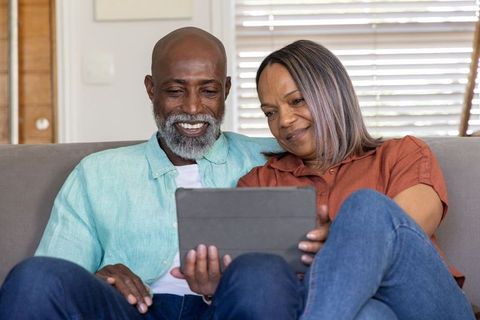 Mature black couple relaxing on sofa sharing tablet and smiling, cozy home moment