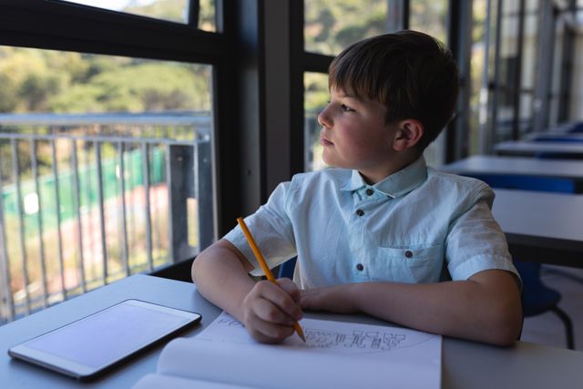 Schoolboy sitting at desk in classroom drawing in notebook while looking away thoughtfully. Ideal for educational content, school-related articles, and creativity-focused materials.