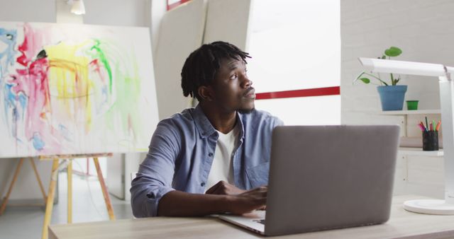 Thoughtful african american male artist with laptop sitting on his desk at art studio. art, hobby and creative occupation concept