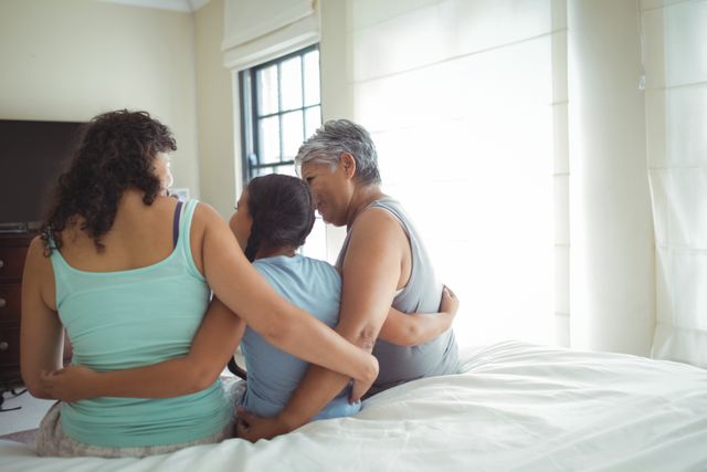 Three women from different generations are sitting on a bed, embracing each other. The scene captures a moment of togetherness and affection, highlighting the bond between family members. This image can be used for themes related to family, love, support, and multi-generational relationships. Ideal for advertisements, blogs, or articles focusing on family values, home life, and emotional connections.