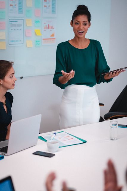 This image shows a confident biracial businesswoman standing and talking to her colleagues during an office meeting. Ideal for use in articles or presentations about leadership, teamwork, corporate culture, business strategy, and professional environments.