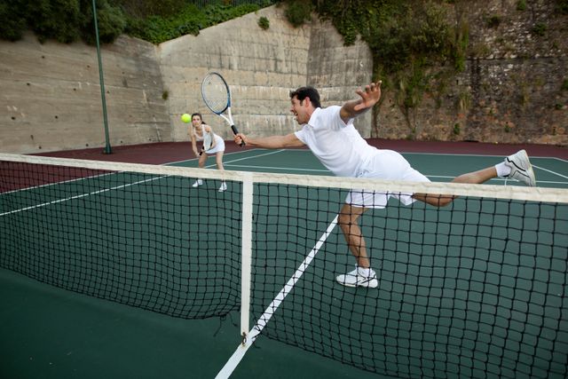 Caucasian man and woman engaged in a tennis match on an outdoor court. The man is hitting the ball while the woman stands in the background. Ideal for use in sports-related content, fitness promotions, and advertisements for athletic gear or tennis equipment.