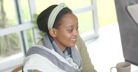 African American woman smiling and chatting over coffee by window, wearing scarf and headband