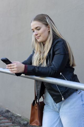 Young woman leaning on railing scrolling phone in leather jacket urban casual lifestyle