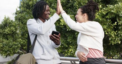 Young friends high-fiving while checking smartphone outdoors, laughing casual urban lifestyle