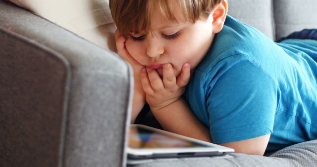 A young Caucasian boy lies on a couch looking at a tablet screen, with copy space. His expression and body language suggest he is engaged or bored with the content he's viewing.