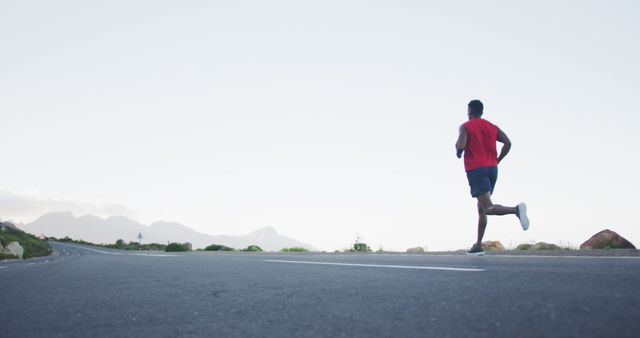 Man running on an empty road surrounded by mountains in the early morning. Man is wearing athletic gear and enjoying an outdoor workout. Suitable for promoting fitness, sportswear, healthy living, or outdoor activities.