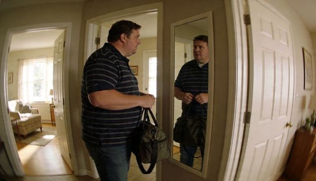 Man standing in home hallway facing floor mirror holding duffel bag wearing striped polo