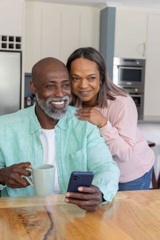 Mature couple sharing smartphone and coffee at modern kitchen table, smiling and relaxed