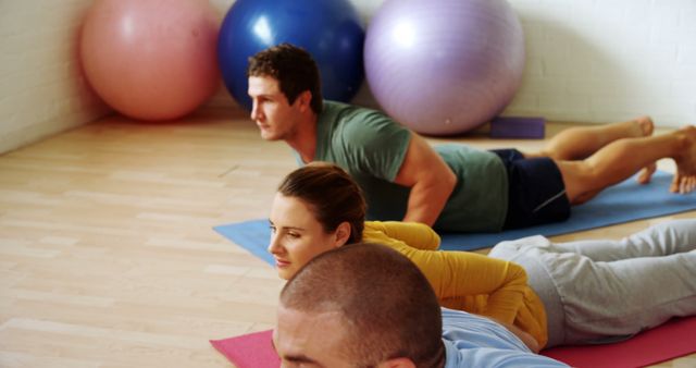 Group of people practicing yoga indoors in a fitness studio, performing Cobra Pose on yoga mats. Exercise balls are visible in the background. Ideal for content related to wellness, group fitness, yoga practice, health awareness, and workout routines.