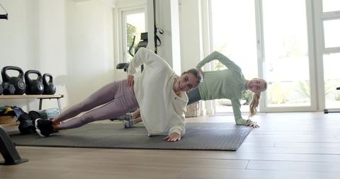 Two women holding side plank on mats in bright home gym with kettlebells and smiles