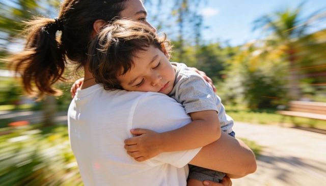 Mother carrying sleeping toddler on shoulder in sunlit park, tender parent-child bond