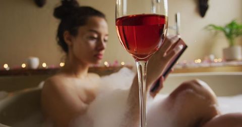 A relaxing scene depicting a woman enjoying a foam bath while reading and sipping on a glass of wine. The soft, ambient lighting enhances the mood of leisure and self-care. This image is perfect for promoting spa products, lifestyle blogs, relaxation articles, or skincare brands focusing on mental wellness and self-indulgence.