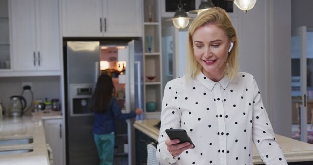 Happy caucasian mother using smartphone and daughter at fridge in kitchen. Family, communication, technology, togetherness and domestic life, unaltered.