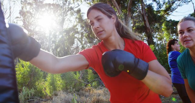 A group of women practicing boxing with a trainer in an outdoor forest setting. One woman is shown throwing a punch towards a pad held by the trainer. The sunlight filtering through the trees adds a natural brightness to the scene. This is ideal for use in materials promoting fitness, outdoor activities, female empowerment, and physical training programs.