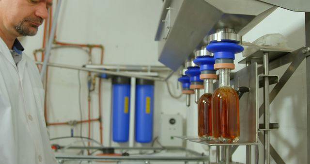 Man supervising automated bottling process in industrial facility. Bottles being filled on production line. This photo can be used in articles about manufacturing, industry practices, modern automation in production, and quality control in factories.