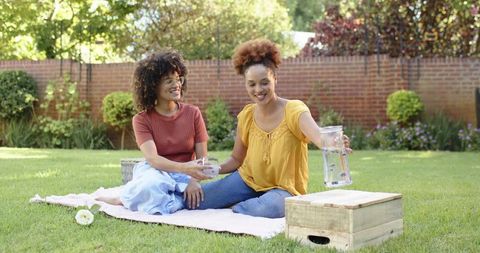 African american women friends sitting on blanket pouring berry-infused water at backyard picnic