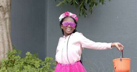 Joyful african american girl wearing flower crown, pink tutu and sunglasses holding pumpkin bucket
