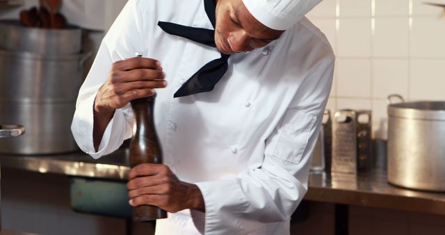 This image shows a chef in a professional kitchen grinding pepper with a spice grinder. The chef is wearing a traditional white uniform and chef hat, indicating a focus on culinary arts and food preparation. Ideal for use in content related to cooking, culinary education, food seasoning, restaurant promotions, and gourmet cuisine.