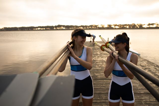 Front view of two Caucasian female rowers wearing sportswear training on the river, standing on a jetty holding oars on their shoulders and looking at each other and smiling.