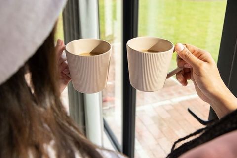 Two women holding matching beige ribbed mugs by sunlit sliding glass door morning tea