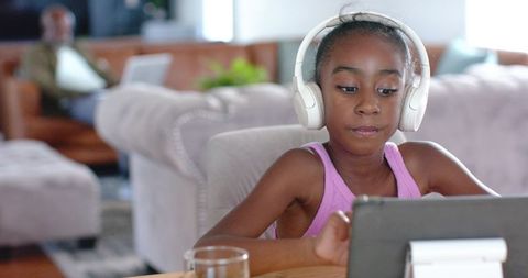 African american girl wearing headphones using tablet for remote learning with father nearby