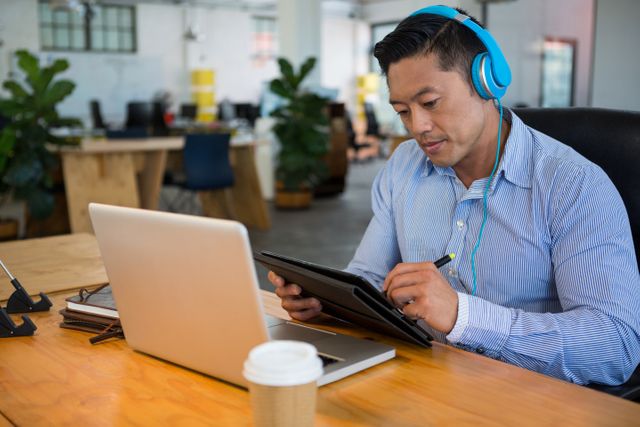Graphic designer using a graphic tablet and laptop in a modern office environment. Wearing headphones and casual attire, he is focused on his work. Ideal for use in articles or advertisements related to creative professions, digital art, modern workspaces, and technology in the workplace.