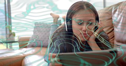 Asian teen girl lounging on leather sofa listening with headphones and using tablet