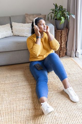 Woman sitting on jute rug listening to headphones in cozy sunlit living room mustard sweater