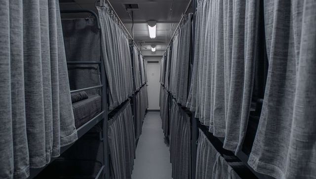 Narrow dormitory corridor featuring stacked metal bunks lined with privacy curtains
