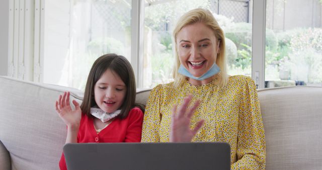 Mother and daughter using a laptop for a video call, both smiling and waving. The setting appears to be a comfortable living room with natural light coming in. Ideal for use in contexts showcasing family bonding, use of technology for remote communication, and home activities during the daytime.