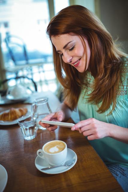 Woman smiling while taking a photo of her latte with a smartphone in a café. Ideal for themes related to social media, lifestyle, food photography, and coffee culture. Use in blogs, articles, or websites focusing on café trends, technology usage in casual settings, or everyday moments.